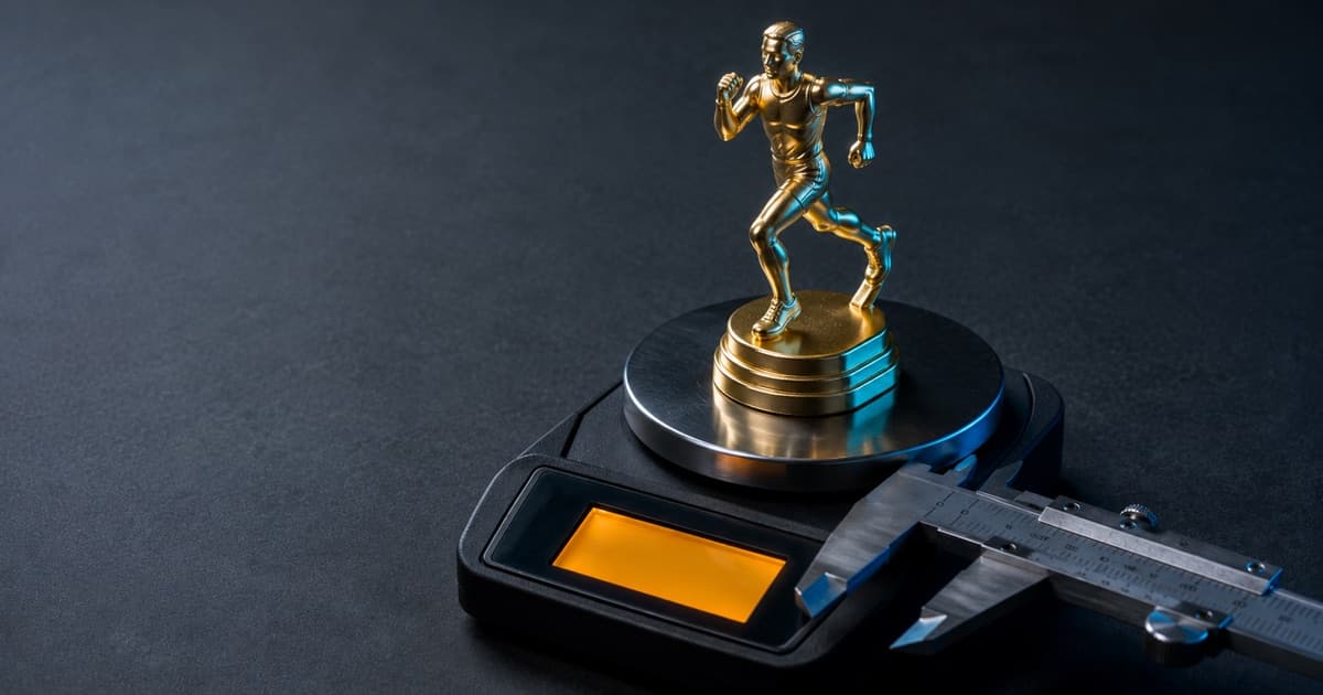 A gold plastic running-figure trophy standing on the steel platter of a precision laboratory balance, machinist's calipers open beside it and the balance readout glowing amber, on a dark editorial surface lit with cool blue rim light.