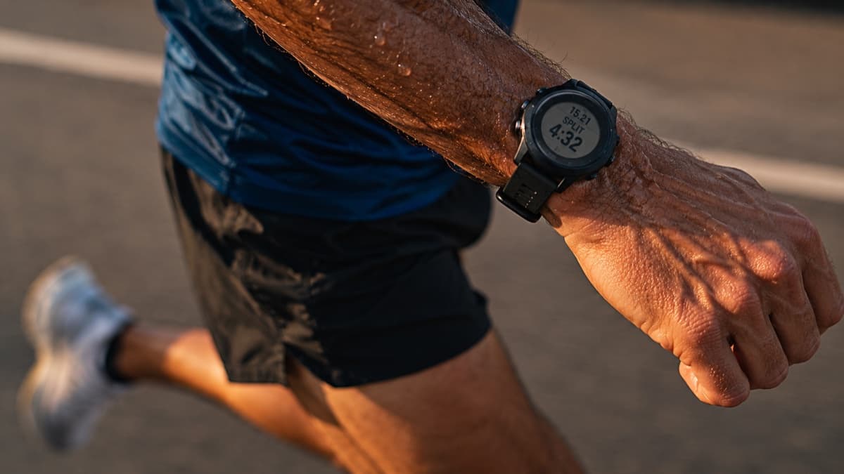 Close-up of a masters runner's forearm and GPS watch mid-race, sweat visible, blurred road in the warm background.